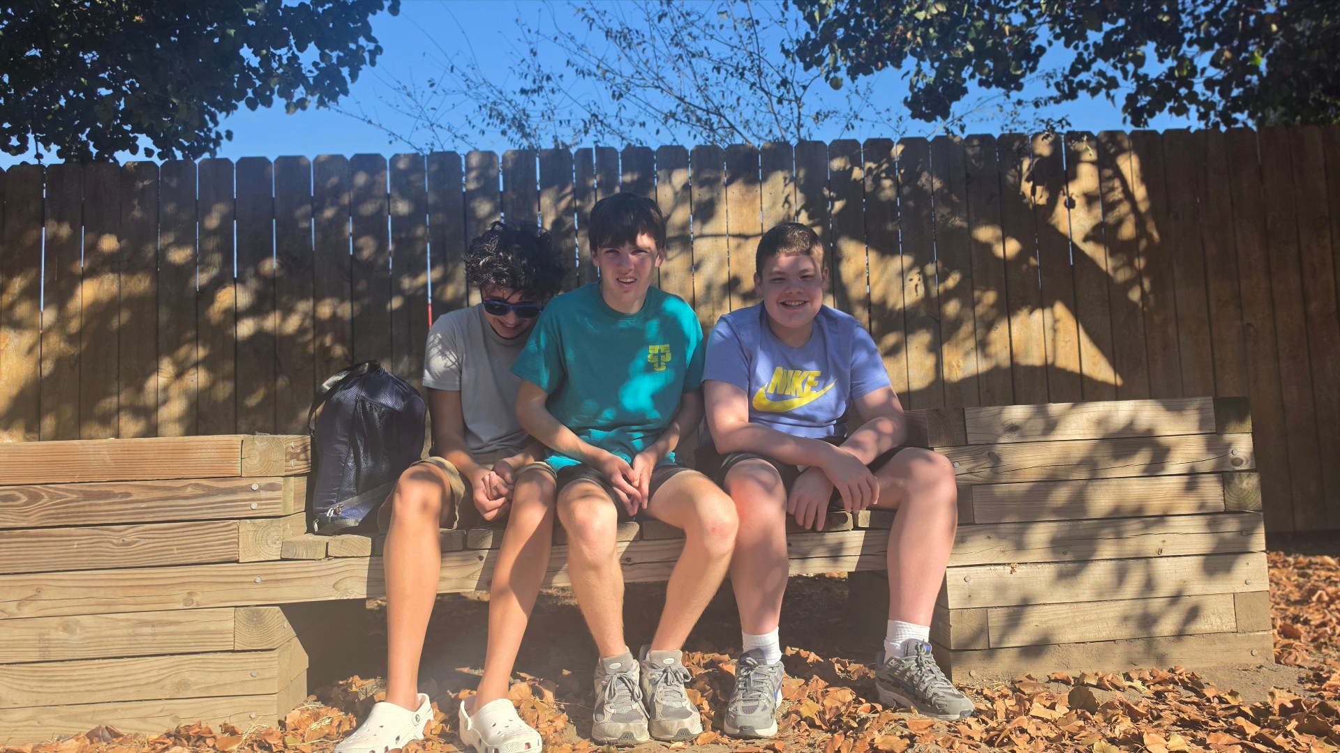 Three young people sit together on a wooden bench outdoors.